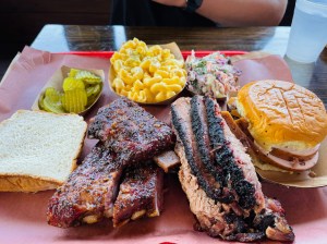 A large platter of barbecue meats and sides, including pork ribs, beef brisket, smoked turkey, mac and cheese, cole slaw, pickles, and bread.