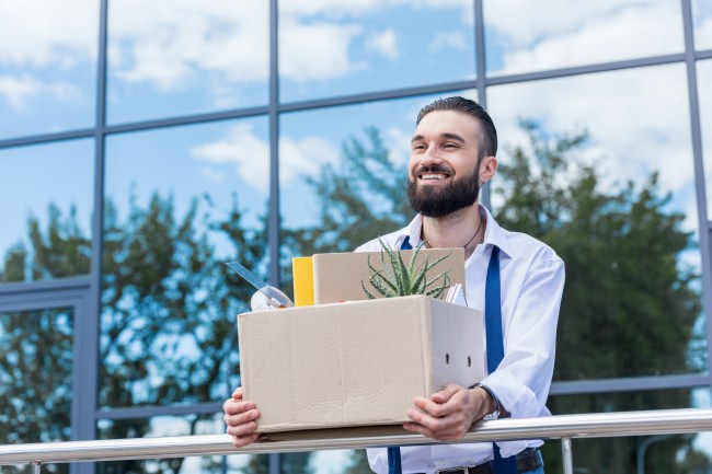 happy businessman with cardboard box with office supplies in hands standing outside office building, quitting job concept
