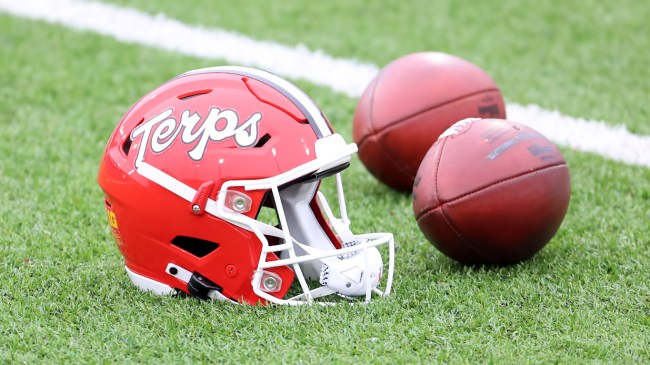 A Maryland Terrapins helmet sits on the field next to a pair of footballs.