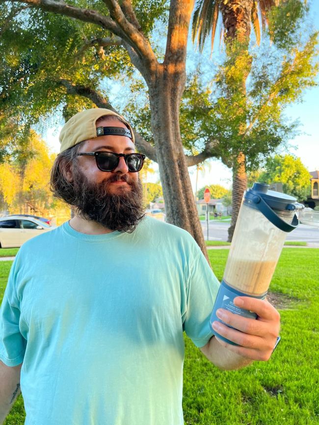 Beard man in sunglasses and a backwards hat holding a Ninja Blast Portable Blender in a city park after making a delicious peach smoothie with it.